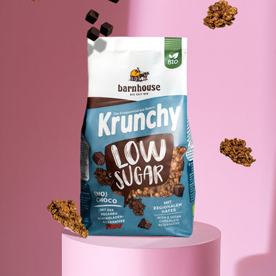 Blick von vorne auf einen blau-braun-weißen Beutel Krunchy Low Sugar (No) Choco von Bio-Knuspermüsli-Hersteller Barnhouse, der auf einer rosefarbenen Sockel steht. Außenrum schweben Krunchy-Cluster sowie Schokowürfel der veganen Schoko-Alternative THIC. Der Hintergrund ist ebenfalls rosa.