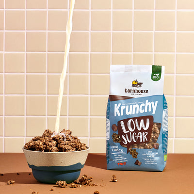 Blick von vorne auf einen blau-braun-weißen Beutel Krunchy Low Sugar (No) Choco von Bio-Knuspermüsli-Hersteller Barnhouse, der auf einer braunen Ablage steht. Links daneben eine blau-graue Schüssel mit diesem Krunchy, in die von oben ein Milchstrahl fließt. Außenrum liegen Krunchy-Cluster. Die Wand im Hintergrund ist weiß-beige gefliest.