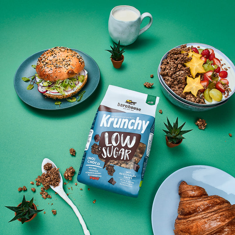 Blick von oben auf eine grüne Ablage, auf der ein blau-braun-weißer Beutel Krunchy Low Sugar (No) Choco von Bio-Knuspermüsli-Hersteller Barnhouse liegt. Außenrum stehen 3 Kakteen, ein Löffel mit Krunchy-Clustern sowie eine Schüssel mit diesem Müsli, Sternenfrucht, Weintrauben, Erdbeeren und Milch. Zudem stehen noch eine Tasse mit Milch, eine Teller mit Croissant sowie ein Teller mit einem Bagel auf dem Tisch.