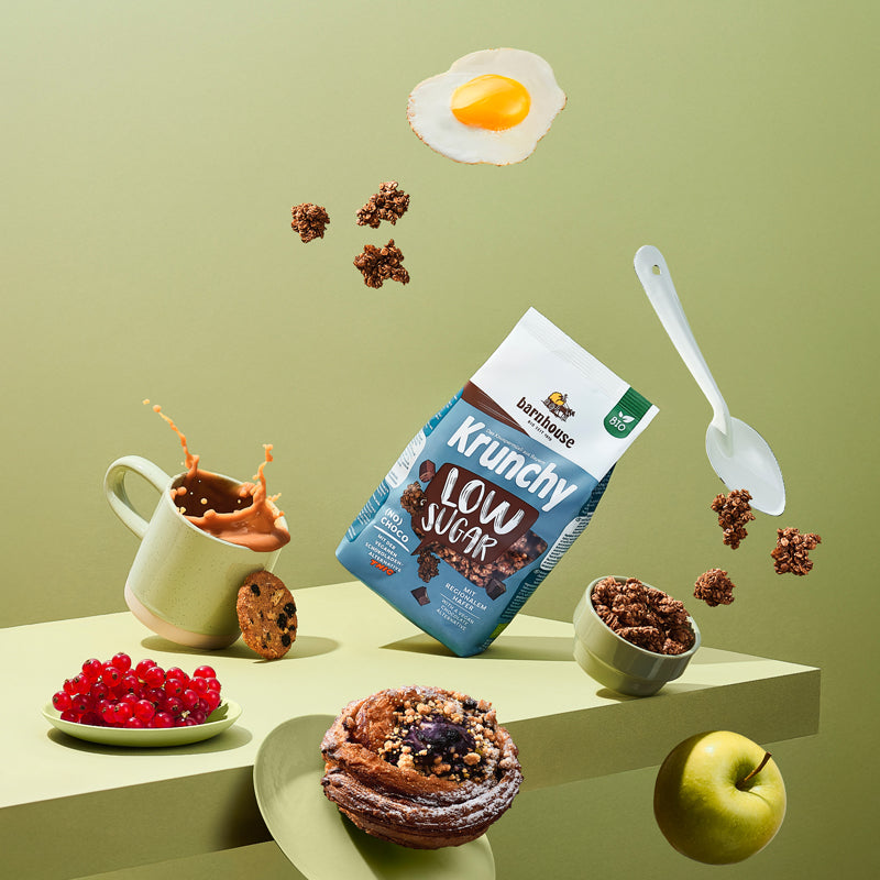 Blick voller Schwung von vorne auf ein grünes Holzbrett. Alle Elemente auf dem Bild fliegen durch die Luft: Braun-blau-weißer Beutel Krunchy Low Sugar (No) Choco von Bio-Knuspermüsli-Hersteller Barnhouse, Schale mit diesem Müsli, Krunchy-Cluster, weißer Löffel, Spiegelei, Tasse mit Kaffee, Schoko-Keks, Teller mit Johannisbeeren, Teller mit Nussschnecke und ein grüner Apfel.