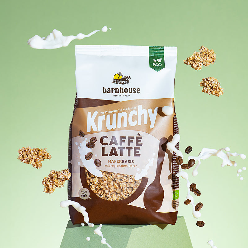 Braun-beiger Beutel Krunchy Caffè Latte von Bio-Knuspermüsli-Hersteller Barnhouse, der auf einem grünen Sockel vor einem grün-weißen Hintergrund steht. Außemrum schweben ein paar Cluster sowie Kaffeebohnen herum. Außerdem sind unten ein paar Milch-Spritzer zu sehen - passend zur Packung. Und oben schwebt ein Milchstrahl in Form einer Wolke.
