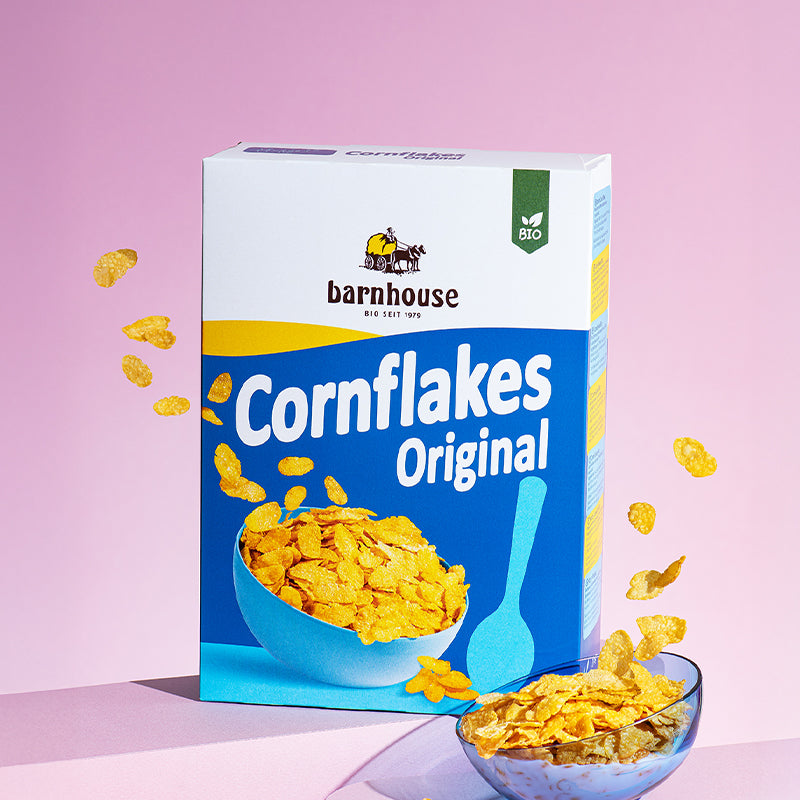 Blauer Karton Cornflakes Original von Bio-Knuspermüsli-Hersteller Barnhouse, der vor einem rosa Hintergrund auf einer rosa Erhöhung steht. Außemrum schweben ein paar Cornflakes rum.  Vorne rechts ist noch eine dunkelblaue, durchsichtige Schüssel zu sehen, die ganz leicht nach links geneigt und mit Cornflakes sowie Milch befüllt ist.