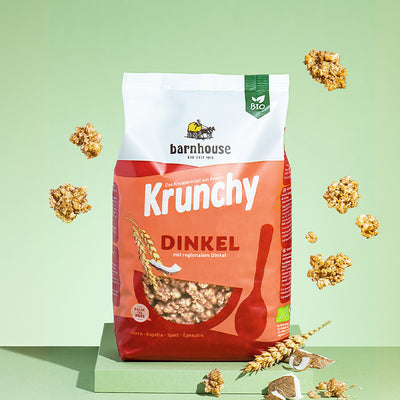 Beutel Krunchy Dinkel von Bio-Knuspermüsli-Hersteller Barnhouse, der auf einer grünen Platte steht. Von oben fallen Knuspermüsli-Cluster vor grünem Hintergrund nach unten. Um den Beutel herum liegen noch weitere Cluster, Stücke einer Kokosnuss sowie ein Dinkelhalm.