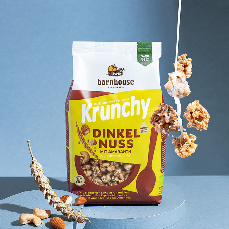 Beutel Krunchy Dinkel-Nuss mit Amaranth von Bio-Knuspermüsli-Hersteller Barnhouse, der auf einem runden, blauen Podest steht, das auf einem blauen Untergrund platziert ist. Von oben fallen Knuspermüsli-Cluster nach unten, in die ein Milchstrahl fließt. Links ist ein weiteres, blaues Podest mit Dinkelhalm, Cashews und Mandeln.