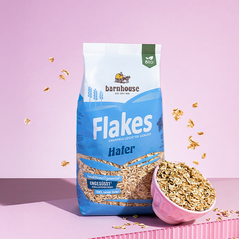 Blauer Beutel Flakes Hafer von Bio-Knuspermüsli-Hersteller Barnhouse, der vor einem rosa Hintergrund auf einer rosa Erhöhung steht. Im ganzen Bild schweben Haferflakes rum.  Vorne rechts ist noch eine rosa Schüssel zu sehen, die ca. 45 Grad nach vorne geneigt und mit Flakes Hafer gefüllt ist.