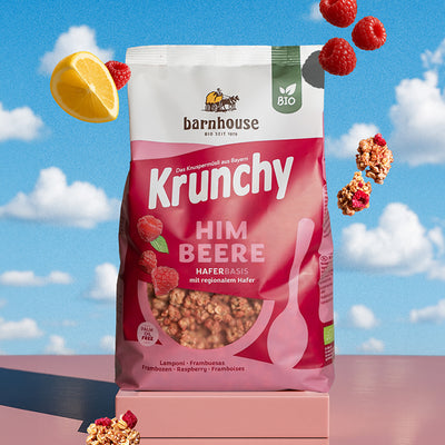 Beerenfarbiger, rosa-weißer Beutel Krunchy Himbeere von Bio-Knuspermüsli-Hersteller Barnhouse auf einem eckigen Sockel. Von oben fallen Knuspermüsli-Cluster, Himbeeren & 1 Zitronen-Viertel vor einem Hintergrund mit blauem Himmel und Wolken nach unten.