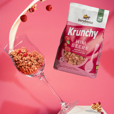 Beutel Krunchy Himbeere, der rechts neben einem Weinglas schwebt, das links von einem weißen Sockel fällt. Das Weinglas ist mit Clustern des Himbeer-Müslis befüllt. Vom oberen Bildrand fließt ein Milchstrahl ins Glas und weitere Cluster sowie Himbeeren fliegen herum.