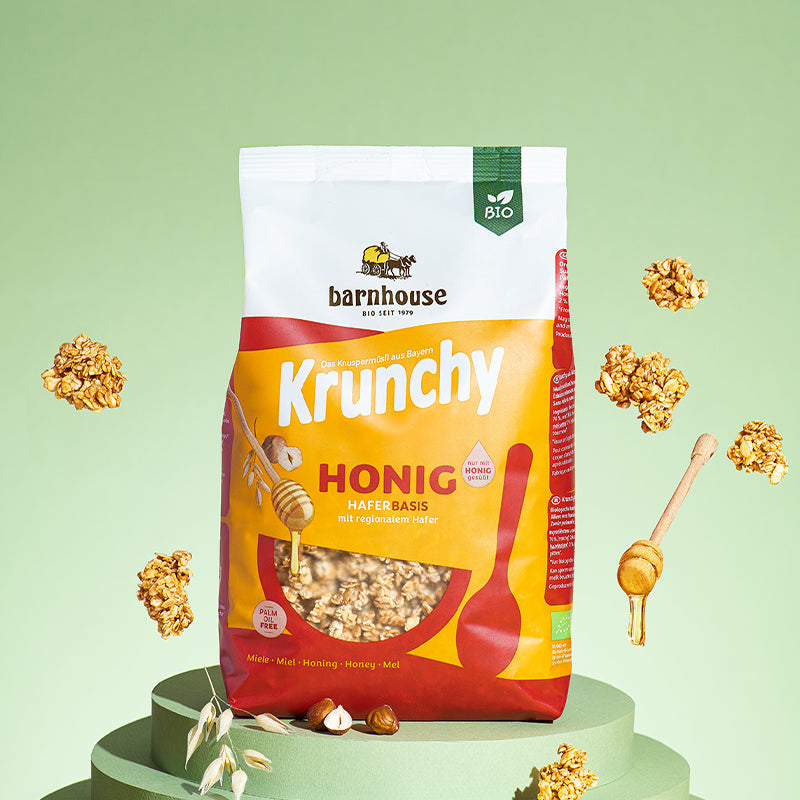 Beutel Krunchy Honig von Bio-Knuspermüsli-Hersteller Barnhouse, der auf 3 runden Scheiben steht, die nach unten hin immer breiter werden. Von oben fallen Knuspermüsli-Cluster sowie ein Honiglöffel mit Honig vor einem grünen Hintergrund nach unten. Auf den grünen Scheiben liegen noch Haselnüsse und Haferknospen, die Zutaten des Honig-Müslis sind.
