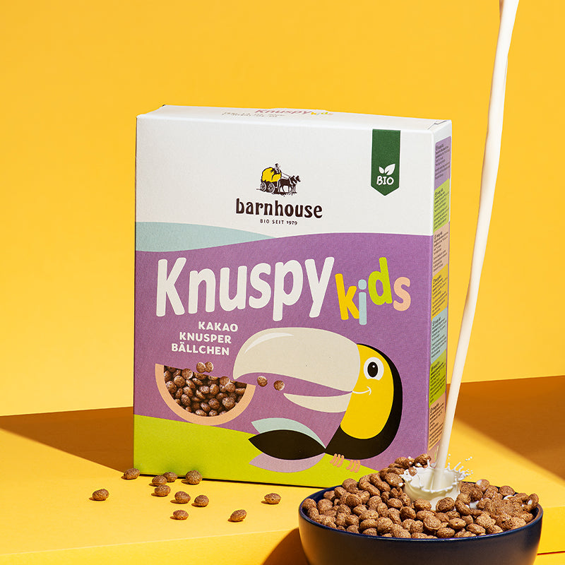 Violetter Karton Knuspy Kids von Bio-Knuspermüsli-Hersteller Barnhouse, der vor einem gelben Hintergrund auf einer gelben Erhöhung steht. Vor der Packung liegen ein paar Knuspy Kids und vorne rechts ist eine dunkelblaue Schüssel zu sehen, die mit Knuspy Kids befüllt ist. Vom oberen Bildrand fließt ein Milchstrahl in die Schüssel.