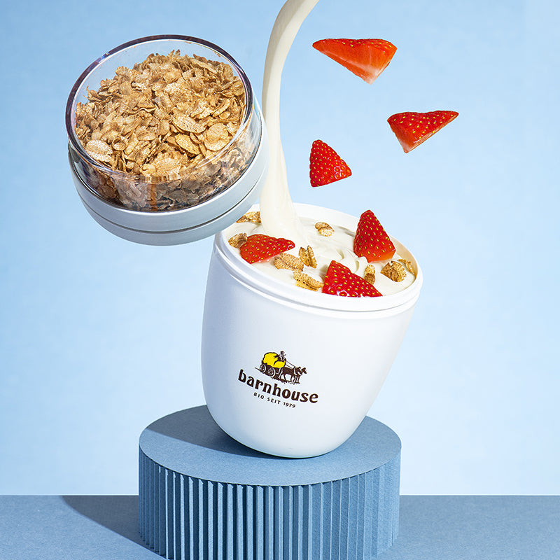 Krunchy2go-Becher von Bio-Knuspermüsli-Hersteller Barnhouse, der schräg auf einem blauen, runden Sockel steht. Von oben fallen Joghurt, Erdbeerstücke und Dinkelflocken vor blauem Hintergrund in den Becher mit Barnhouse Logo. Der abgeschraubte Deckel mit Dinkelflocken fliegt links über dem Becher.