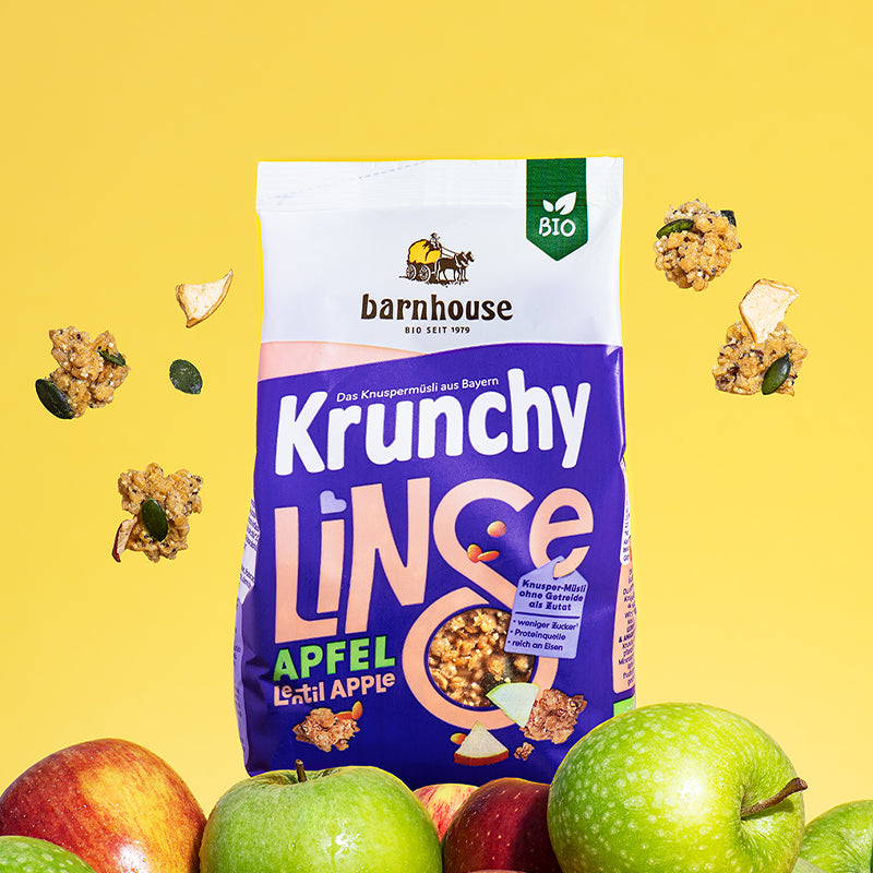 Blau-weißer Beutel Krunchy Linse mit Apfel von Bio-Knuspermüsli-Hersteller Barnhouse, der auf einem Berg aus roten und grünen Äpfeln steht. Von oben fallen Knuspermüsli-Cluster mit Apfelstücken und Sonnenblumenkernen vor einem gelben Hintergrund nach unten.