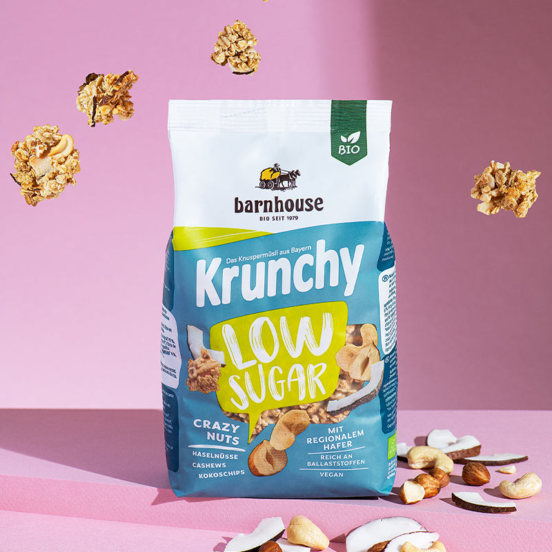 Grün-weiß-blauer Beutel Krunchy Low Sugar Crazy Nuts von Bio-Knuspermüsli-Hersteller Barnhouse, der auf einem rosa Untergrund steht. Von oben fallen Knuspermüsli-Cluster vor einem rosa Hintergrund nach unten. Am Boden liegen Haselnüsse, Cashewnüsse und Kokoschips - die Zutaten des nussigen, zuckerarmen Müslis.