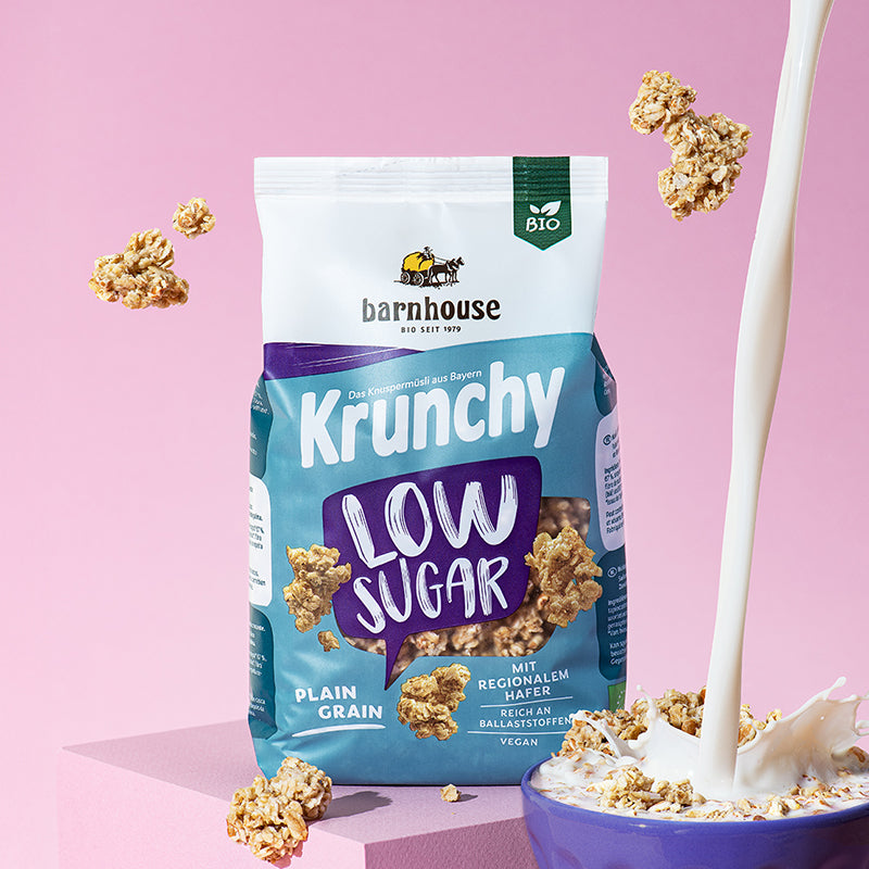 Lila-weiß-blauer Beutel Krunchy Low Sugar Plain Grain von Bio-Knuspermüsli-Hersteller Barnhouse, der auf einem rosa Untergrund steht. Von oben fallen Knuspermüsli-Cluster vor einem rosa Hintergrund nach unten. Vorne rechts steht eine dunkelblaue Schale mit dem zuckerarmen Müsli, in die von oben ein Milchstrahl reingeschüttet wird.