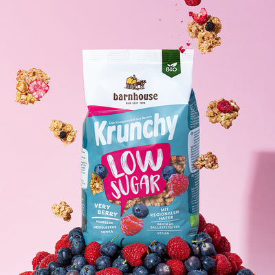Pink-weiß-blauer Beutel Krunchy Low Sugar Very Berry von Bio-Knuspermüsli-Hersteller Barnhouse, der auf einem Berg aus Himbeeren und Heidelbeeren steht. Von oben fallen Knuspermüsli-Cluster mit den Beeren vor einem rosa Hintergrund nach unten.