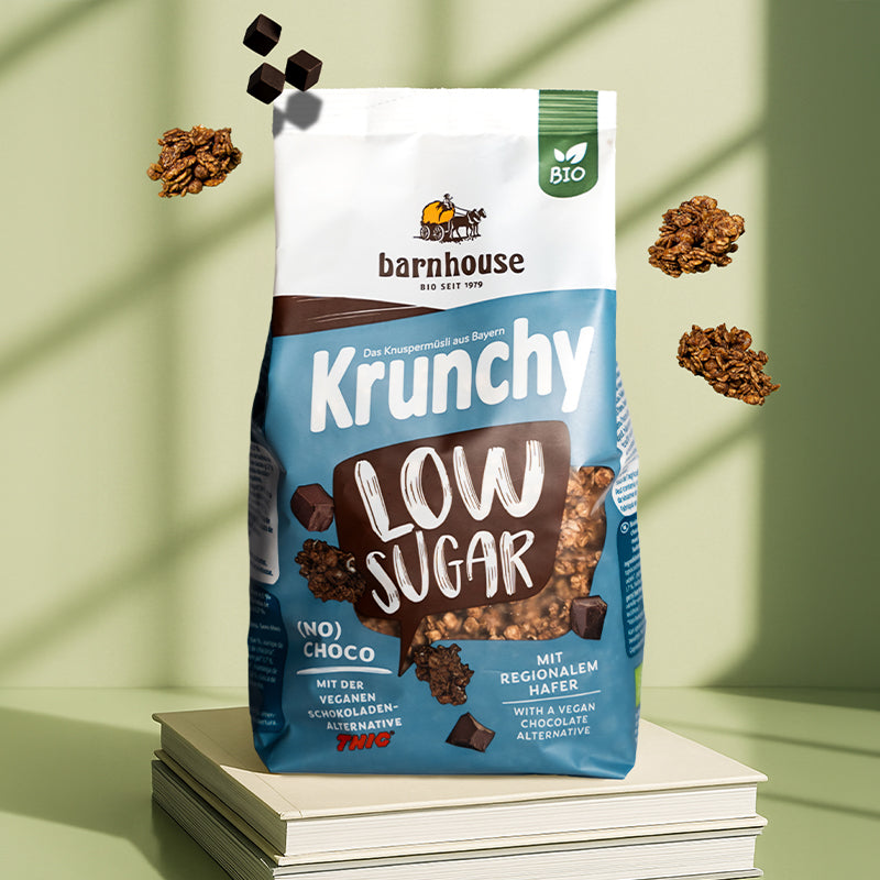 Blau-braun-weißer Beutel Krunchy Low Suagr (No) Choco von Bio-Knuspermüsli-Hersteller Barnhouse, der auf 2 hellen Büchern steht. Von oben fallen Knuspermüsli-Cluster und THIC-Schoko-Stücke vor einem grünen Hintergrund nach unten. Von außen strahlt Sonne durch ein Fenster in den Raum.
