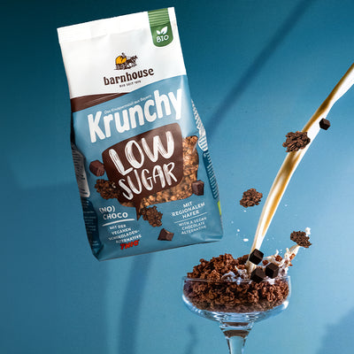 Beutel Krunchy Low Sugar (No) Choco, der über einem Glas schwebt, das mit Knuspermüsli-Clustern befüllt ist. Vom rechten Bildrand fließt ein Milchstrahl ins Glas und "Schoko"-Cluster fliegen herum.