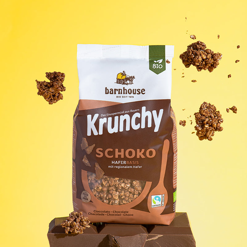 Braun-weißer Beutel Krunchy Schoko von Bio-Knuspermüsli-Hersteller Barnhouse, der auf einer dicken Tafel Schokolade steht. Von oben fallen Knuspermüsli-Cluster vor einem gelben Hintergrund nach unten.
