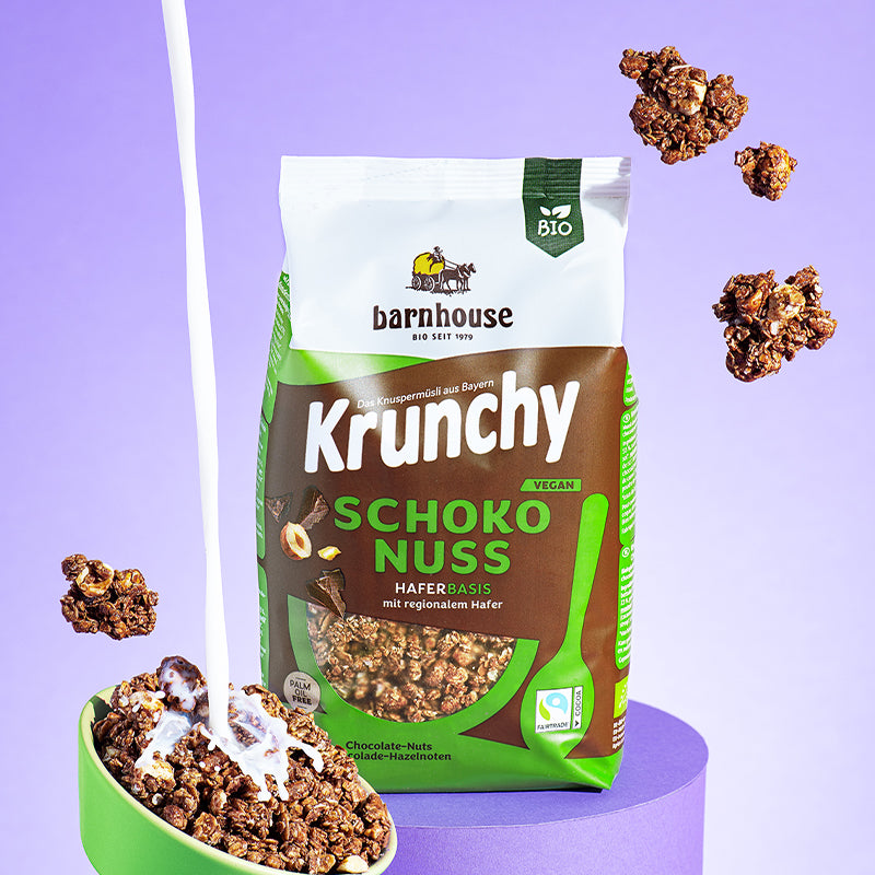 Grün-weiß-brauner Beutel Krunchy Schoko-Nuss von Bio-Knuspermüsli-Hersteller Barnhouse, der auf einem violetten, runden Sockel steht. Von oben fallen Knuspermüsli-Cluster vor einem violetten Hintergrund nach unten. Vorne links steht sehr schräg eine grüne Schale mit dem Schoko-Müsli, in die von oben ein Milchstrahl reingeschüttet wird.