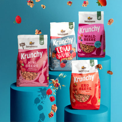 Blick von vorne auf die Beutel der vier Frucht-Krunchys Himbeere, Low Sugar Very Berry, Waldbeere weniger Zucker und Erdbeere von Bio-Knuspermüsli-Hersteller Barnhouse. Die Packungen stehen jeweils auf einem runden, blauen Sockel. Von oben fallen Knuspermüsli-Cluster, ein Haferhalm sowie Erdbeeren, Himbeeren und Heidelbeeren vor blauem Hintergrund nach unten.