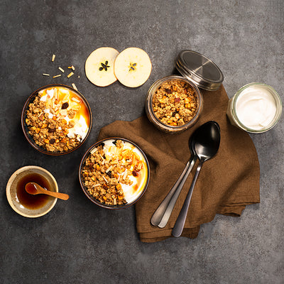 Dunkler Tisch mit einem braunen Geschirrtuch, auf dem 3 Löffel liegen. Daneben befindet sich ein durchsichtiges Gefäß mit Krunchy Apfel-Zimt von Bio-Knuspermüsli-Hersteller Barnhouse. Zudem 2 Schüsseln, die jeweils mit Krunchy Apfel-Zimt, Joghurt und Apfeldicksaft befüllt sind. Zudem stehen noch 2 Apfelscheiben, eine Schüssel mit Apfeldicksaft und Holzlöffel sowie eine Glas mit Joghurt aus dem Tisch.