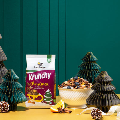 Beutel Krunchy Christmas von Bio-Knuspermüsli-Hersteller Barnhouse, der auf einem gelben Untergrund vor einer grünen Wand steht. Für die weihnachtliche Deko sorgen kleine, künstliche Christbäume, Tannenzapfen, eine Orangenscheibe und Zimtstange. In der Mitte steht noch eine weiße Schüssel, die mit unserem Weihnachts-Krunchy bis oben hin gefüllt und mit Heidelbeeren dekoriert ist.