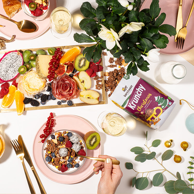 Bunt dekorierter Tisch vom Weihnachts-Brunch mit Obst, Wurst und Co. Edles, goldenes Besteck, goldene Weihnachtskugeln und weiß-grüne Blumen sorgen für noch schöneres Ambiente. In der Mitte liegt ein Beutel Krunchy Christmas von Bio-Knuspermüsli-Hersteller Barnhouse, aus dem Cluster auf den Tisch rieseln.