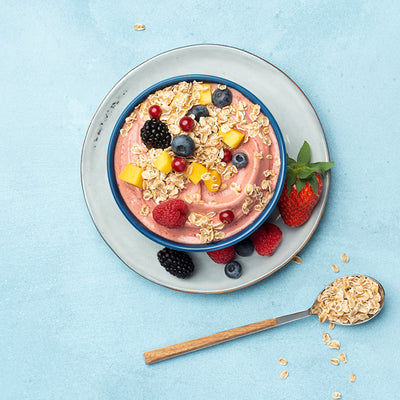 Hellblauer Untergrund, auf dem ein weiß-grauer Teller steht. Auf diesem Teller befindet sich eine dunkelblaue Schüssel, die mit rötlichem Joghurt, jeder Menge Beeren und Mango befüllt ist. Auch auf dem Unterteller befinden sich noch verschiedene Beeren. Als Topping sind noch Flakes Hafer von Bio-Knuspermüsli-Hersteller Barnhouse mit drauf.. Neben der Schüssel befindet sich ein grau-brauner Löffel, der mit Haferflakes gefüllt ist. Daneben liegen noch weitere Haferflakes.