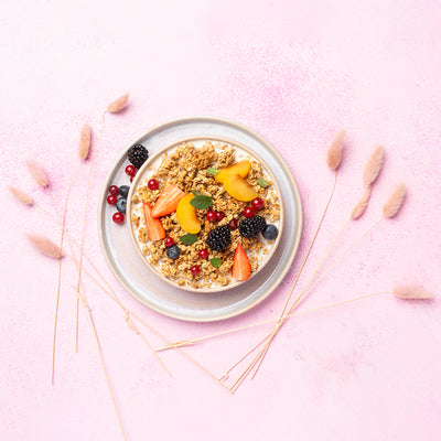 Rosa Untergrund mit flauschigen Blumen, auf dem ein weißer Unterteller mit Johannisbeeren, Heidelbeeren und einer Brombeere steht. Darauf befindet sich eine Schüssel mit Milch, Krunchy Honig, frischen Erdbeerstücken, Aprikosen, Johannisbeeren, Heidelbeeren, Brombeeren und etwas Minze.