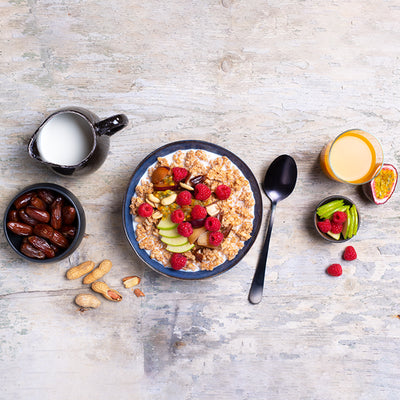 Ein heller Holztisch, in dessen Mitte eine blaue Müslischale steht, die mit Milch & Krunchy Klassik Hafer von Bio-Knuspermüsli-Hersteller Barnhouse befüllt ist. Als Topping liegen Himbeeren, Apfelscheiben, Nüsse, Pflaumen, Datteln und eine Passionsfrucht schön angerichtet obendrauf. Rechts daneben sind ein Löffel, ein Glas Orangensaft, eine halbe Passionsfrucht, eine Schüssel mit Apfelscheiben und Himbeeren. Links steht eine schwarze Kanne mit Milch, eine schwarze Schale mit Datteln und Erdnüsse liegen da.