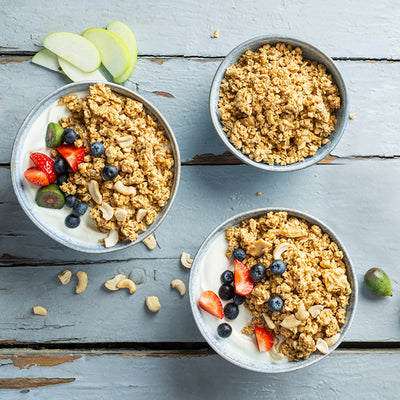 Blick von oben auf einen Holztisch, auf dem ein paar grüne Apfelspalten, eine Erdbeere, eine Minikiwi und Cashewnüsse liegen. Oben rechts steht eine blaue Müslischüssel mit Krunchy Low Sugar Crazy Nuts von Bio-Knuspermüsli-Hersteller Barnhouse. Im unteren Bereich steht eine weitere blaue Schale mit dem zuckerarmen Nuss-Müsli, allerdings mit Joghurt und Erdbeeren sowie Heidelbeeren als Topping. Links steht noch eine blaue Müslischale, die zudem noch mit zwei halben Minikiwis dekoriert ist.