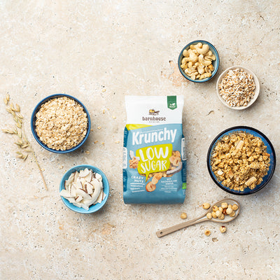 Blick von oben auf einen grün-weiß-blauen Beutel Krunchy Low Sugar Crazy Nuts von Bio-Knuspermüsli-Hersteller Barnhouse auf einer beigen Marmorplatte. Außenrum stehen vier verschiedenfarbige Schalen mit den Zutaten Kokoschips, Cashewnüssen, gepuffter Dinkel & Haferflocken sowie ein Löffel mit Haselnüssen. Eine weitere Schale ist mit dem nussigen, zuckerarmen Knuspermüsli befüllt. Ein Haferhalm liegt auch dabei. 