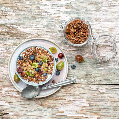 Blick von oben auf einen weiß gestrichenen Holztisch, der schon etwas abblättert. Oben steht ein geöffnetes Vorratsglas mit Krunchy Schoko-Nuss von Bio-Knuspermüsli-Hersteller Barnhouse. Darunter liegen ein Weintraube und eine Heidelbeere. Der Fokus liegt auf der Bowl, die mit dem nussigen Schokoaden-Müsli und Milch befüllt ist. Als Topping liegen Heidelbeeren, weiße Johannisbeeren sowie grüne und rote Stachelbeeren obendrauf. Auf dem Unterteller liegen noch weitere Beeren sowie ein silberner Löffel.