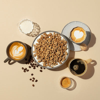 Einfarbiger, beiger Hintergrund, in dessen Fokus eine Schüssel mit Milch und Krunchy Caffè Latte von Bio-Knuspermüsli-Hersteller Barnhouse steht. Außenrum liegen einige Kaffeebohnen. Außerdem stehen dort noch ein Glaskaraffe mit Milch sowie ein Espresso und eine Tasse schwarzer Kaffee. Links steht ein Cappuccino mit Milchschaum in Blumenform in einer grauen Tasse. Auf der rechten Siete steht ein grauer Unterteller mit einem kleinen Keks und einer Cappuccino mit Milchschaum in Herzform.