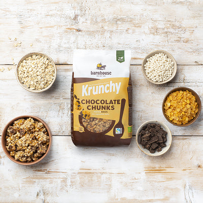 Beutel Krunchy Chocolate Chunks von Bio-Knuspermüsli-Hersteller Barnhouse auf einem weißen Holztisch. Links steht eine braune Schüssel mit diesem Müsli. Zudem stehen dort noch 3 weiße und eine braune Schale, die mit den Zutaten Haferflocken, Cornflakes, Schoko-Stückchen und Knusperreis gefüllt sind.