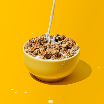 Gelbe Schüssel, die gut gefüllt mit Krunchy Chocolate Banana Chunks von Bio-Knuspermüsli-Hersteller Barnhouse  ist. Vom oberen Bildrand wird ein Strahl Milch hinein geschüttet. Ansonsten sind noch ein paar Milchspritzer auf dem gelben Hintergrund zu sehen.