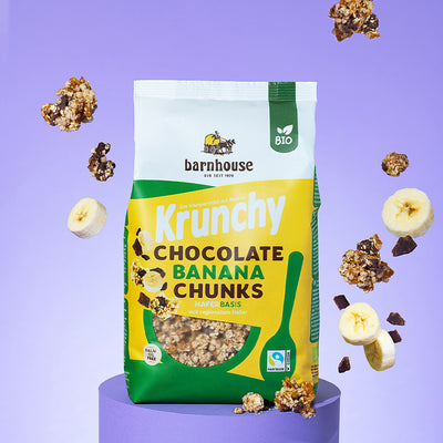 Beutel Krunchy Chocolate Banana Chunks von Bio-Knuspermüsli-Hersteller Barnhouse, der auf einem violetten, runden Socken steht. Von oben fallen Knuspermüsli-Cluster, Bananen- und Schoko-Stücke vor dem violetten Hintergrund nach unten.