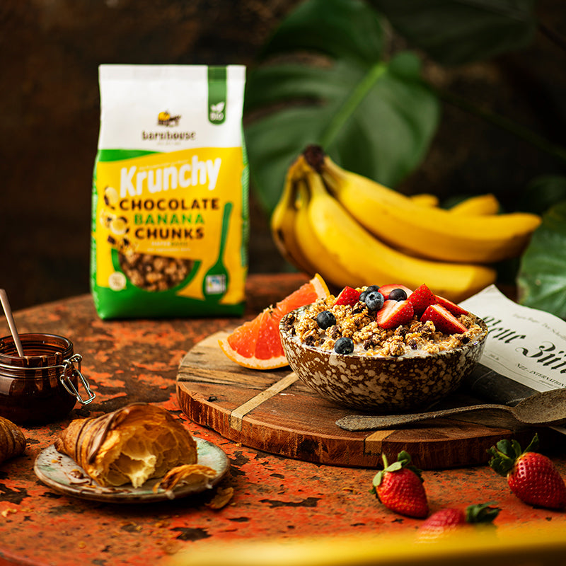 Bunt dekorierter, rötlicher Tisch mit einem Beutel Krunchy Chocolate Banana Chunks von Bio-Knuspermüsli-Hersteller Barnhouse. Außerdem auf dem Tisch: Bananen, ein Schoko-Croissant, Erdbeeren, eine Zeitung, eine Schüssel mit diesem Müsli sowie Erdbeeren und Heidelbeeren, eine Scheibe Blutorange. Im Hintergrund sind die Blätter einer grünen Pflanze zu sehen.