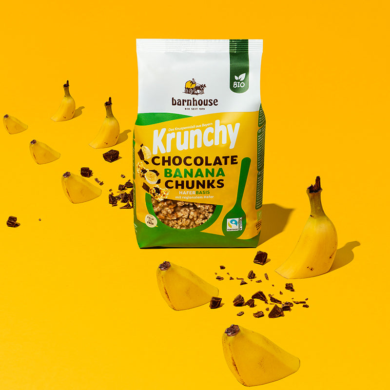 In der Mitte steht ein Beutel Krunchy Chocolate Banana Chunks von Bio-Knuspermüsli-Hersteller Barnhouse. Daneben sind Schoko-Stückchen sowie angeschnittene Bananen zu sehen, die auf dem gelben Boden "sitzen".