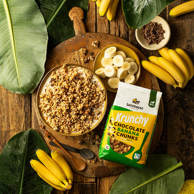 Beutel Krunchy Chocolate Banana Chunks von Bio-Knuspermüsli-Hersteller Barnhouse, der gemeinsam mit einer Schüssel dieses Müslis, paar weiteren Clustern, 2 Löffeln und einer Schale mit Bananenscheiben auf einer Holzplatte liegt. Darunter ein brauner Holztisch, auf dem zudem noch einige Bananen, eine Schale mit Schoko-Stückchen sowie riesige Blätter einer grünen Pflanze liegen.