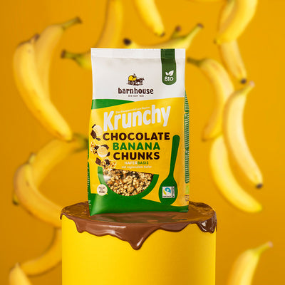 Beutel Krunchy Chocolate Banana Chunks von Bio-Knuspermüsli-Hersteller Barnhouse, der auf einem gelben Sockel steht, dessen Oberfläche mit geschmälzter Schokolade überdeckt ist. Zwischen dem Beutel und dem gelben Hintergrund fallen einige ungeschälte Bananen vom Himmel.