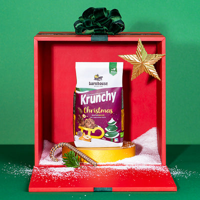 Ein rote, geöffnete Geschenkbox mit goldenem Stern, etwas künstlichem Schnee, einer Zuckerstange, einem Tannenzweig und einer grüner Schleife. Im Geschenk steht ein Beutel Krunchy Christmas von Bio-Knuspermüsli-Hersteller Barnhouse auf einem goldenen Sockel. Die Geschenkbox steht vor einem grünen Hintergrund.