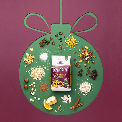 Violetter Hintergrund, in dem die Form einer Christbaumkugel durch grüne Farbe eingezeichnet ist. In diesem Innenraum befindet sich ein Beutel Krunchy Christmas von Bio-Knuspermüsli-Hersteller Barnhouse sowie die einzelnen Zutaten dieses Müslis - Apfelstücke, Orangenscheiben, Haferflocken, Kokoschips, dunkle Schoko-Stückchen Und Co.