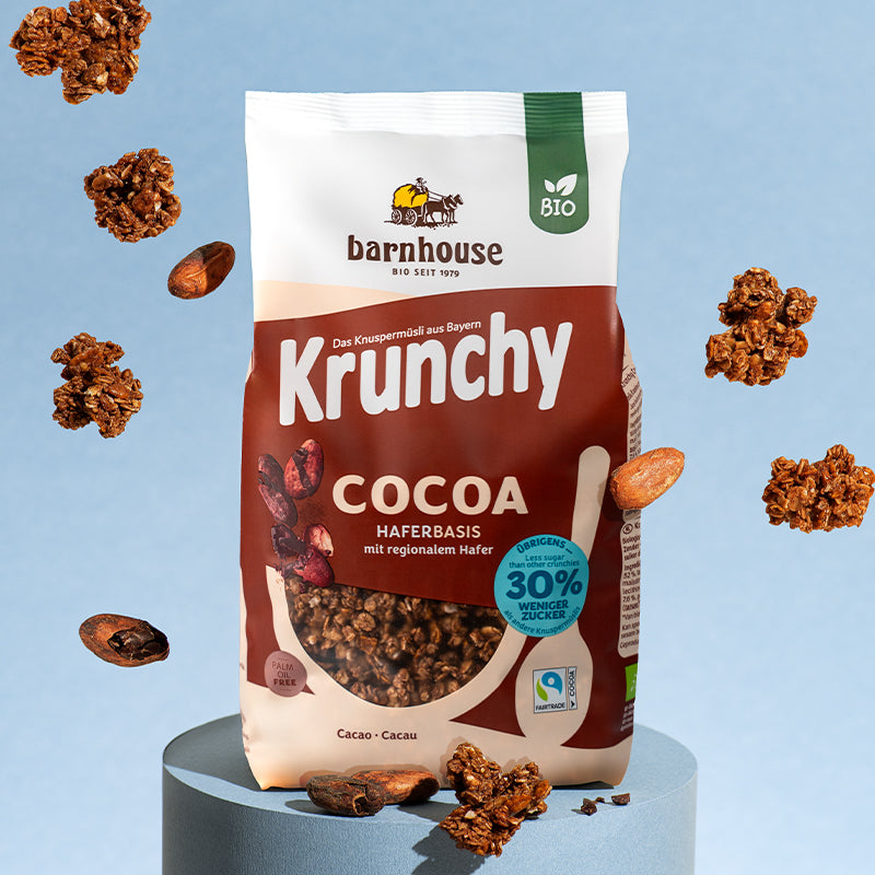 Braun-beiger Beutel Krunchy Cocoa weniger Zucker von Bio-Knuspermüsli-Hersteller Barnhouse auf einem runden, blauen Sockel. Außenrum fliegen Krunchy Cluster und Kakaobohnen.