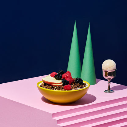 Rosa Treppe, auf der eine gelbe Schüssel mit Krunchy Cocoa weniger Zucker, Milch, Himbeeren, Brombeeren und Apfelscheiben steht. Daneben ein silberner Eierbecher mit Holzkugel sowie 2 dunkelgrüne, hohe Kegel.