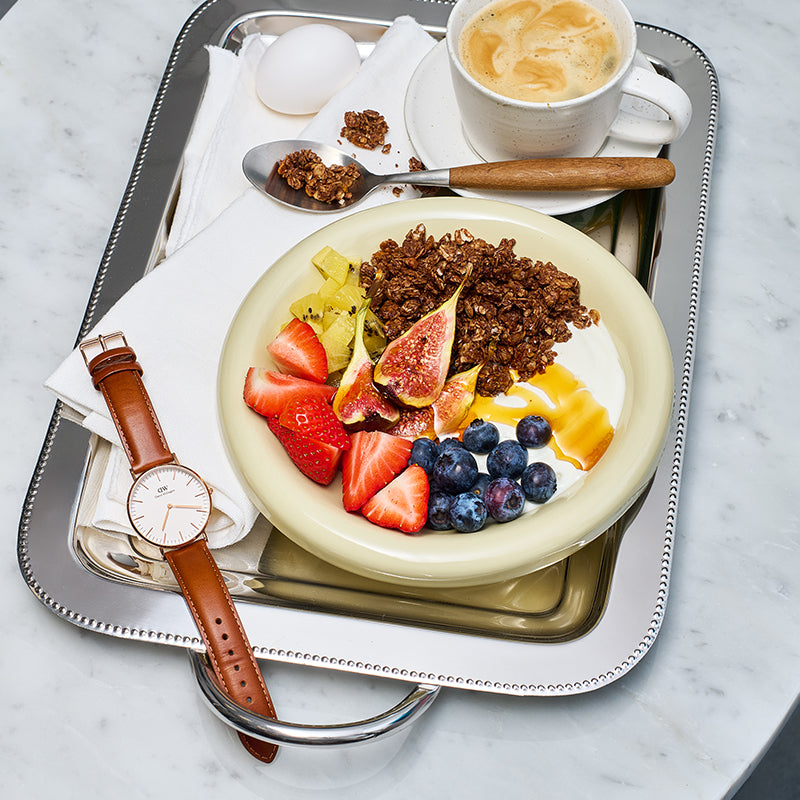 Silbernes Tablett, auf dem eine weiße Stoffserviette, eine braune Uhr, ein Ei, eine Tasse Kaffee sowie ein Löffel mit Clustern von Krunchy Cocoa weniger Zucker liegen. Der Hauptfokus liegt auf einer cremefarbenen Schüssel mit Krunchy Cocoa weniger Zucker, Joghurt, Kiwi, Feigen, Heidelbeeren, Erdbeeren und etwas Honig.