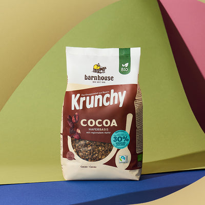 Braun-beiger Beutel Krunchy Cocoa weniger Zucker von Bio-Knuspermüsli Hersteller Barnhouse vor einem bunten Hintergrund mit einer gelben, grünen, roten, blauen und brauen Fläche.
