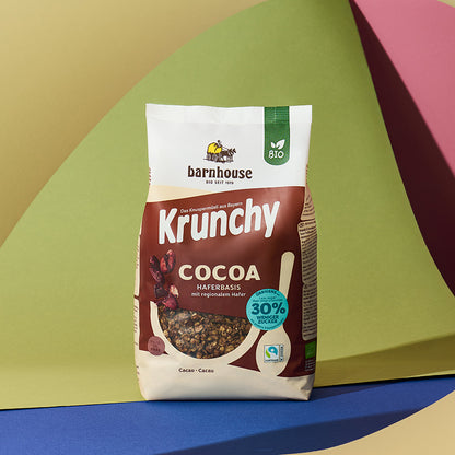 Braun-beiger Beutel Krunchy Cocoa weniger Zucker von Bio-Knuspermüsli Hersteller Barnhouse vor einem bunten Hintergrund mit einer gelben, grünen, roten, blauen und brauen Fläche.