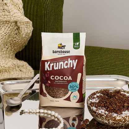 Braun-beiger Beutel Krunchy Cocoa weniger Zucker von Barnhouse, der auf einem silbernen Tablett steht. Links daneben liegen ein silberner Löffel und eine weiße Armkette. Rechts daneben steht eine braun-beige gefleckte Schüssel mit Milch und dem besagten Bio-Knuspermüsli. Das Tablett steht auf einem dunkelgrünen Sessel mit beiger, gehäkelter Decke vor einer weißen Wand.
