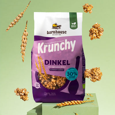 Lila Beutel Krunchy Dinkel weniger Zucker von Barnhouse auf einem grünen, rechteckigen Sockel. Außenrum fliegen Cluster dieses Bio-Knuspermüslis und Dinkelhalme.