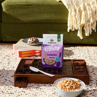 Lila Beutel Krunchy Dinkel weniger Zucker von Barnhouse, der gemeinsam mit einem weißen Löffel auf einem braun-schwarzen Dame-Spielbrett steht. Im Vordergrund steht eine weiße Schüssel mit Milch und dem besagten Bio-Knuspermüsli. Im Hintergrund sind noch eine grüne Couch mit weißer Strickdecke, ein weiß-schwarzer Teppich sowie ein Turm aus einem zusammengeklappten Backgammon-Spielbrett und einem Buch zu sehen, auf dem ein blauer Teller mit einem kleinen Brotlaib steht.